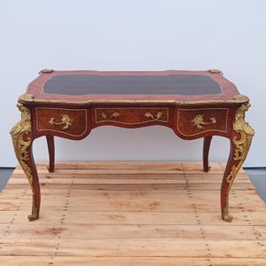 May include: An antique desk with a dark brown leather top and a reddish-brown wooden body. The desk features three drawers with ornate gold-colored handles and decorative gold accents. The legs are curved and also have gold detailing.