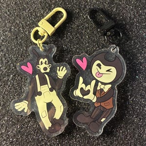 以下が含まれることがあります： 2つの漫画のキャラクターのキーホルダー。1つのキーホルダーは、黄色の衣装と手を振っている白黒のキャラクターが特徴です。もう1つのキーホルダーは、茶色のスーツと舌を出している黒と茶色のキャラクターが特徴です。