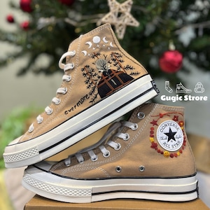 Könnte beinhalten: Ein Paar beigefarbene Converse-Sneaker mit hohem Schaft und individueller Stickerei. Ein Schuh zeigt ein Mond- und Sternenmuster mit dem Wort "evermore", das an der Seite gestickt ist. Der andere Schuh zeigt ein Blumenmuster mit einem roten Stern und dem Converse-Logo.