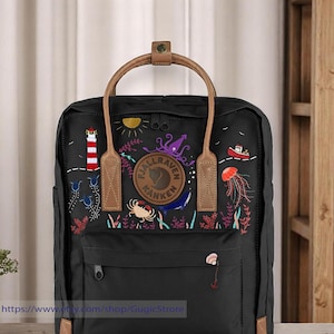 Op de afbeelding: Zwarte Fjällräven Kånken rugzak met bruine leren banden en een kleurrijk geborduurd ontwerp met een vuurtoren, zon, octopus, walvis, kwal en andere zeedieren. Het logo "Fjällräven Kånken" is geborduurd op de voorkant.