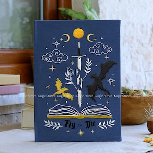 May include: A navy blue book cover with embroidered celestial and fantasy elements. The design includes a sword, clouds, stars, a moon, and the text "Fly or Die". A gold dragon and a black dragon are also featured.