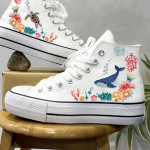 May include: White high-top canvas shoes with colourful embroidered ocean-themed designs, including a whale, coral, and a sea turtle.