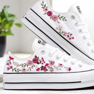 May include: White canvas sneakers with pink and green floral embroidery. The embroidery features pink roses, small pink flowers, and green leaves.