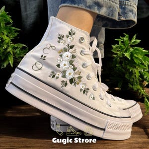 Puede incluir: Zapatillas blancas de lona de caña alta con flores blancas y hojas verdes bordadas. Las zapatillas tienen un ribete negro alrededor de la suela y una puntera de goma negra. Las palabras "&" y "∞" están bordadas en el lateral de la zapatilla.