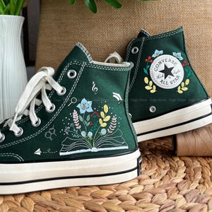 Puede incluir: Zapatillas altas verde oscuro con cordones y suelas blancas. Las zapatillas presentan diseños bordados con motivos florales y científicos, incluyendo un libro abierto, flores y notas musicales. El logo de Converse All Star es visible.