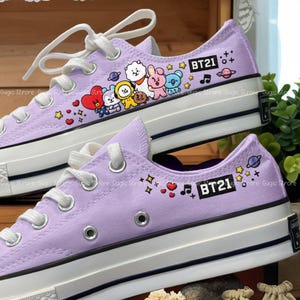 May include: A pair of lavender canvas sneakers with white laces and soles. The sneakers feature colorful cartoon character graphics and the text "BT21". Decorative stars, hearts, and musical notes are also present.