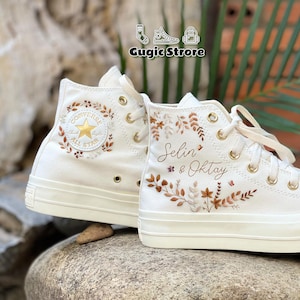 May include: Pair of off-white high-top sneakers with floral embroidery and gold eyelets. The left shoe features the Converse All Star logo. The right shoe is personalized with names and floral designs. The text "Gugic Strore" is at the top.