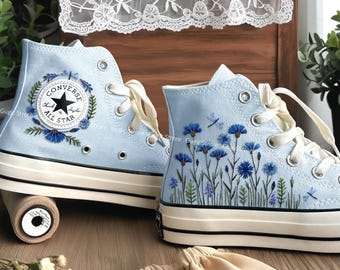May include: Light blue high-top sneakers with embroidered floral and dragonfly designs. One sneaker features the Converse All Star logo. The sneakers have white laces and soles, and are set on a wooden surface.