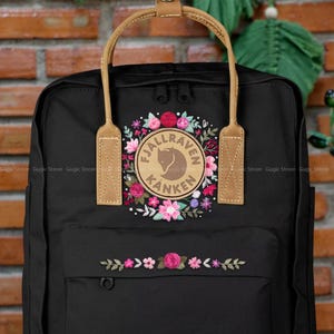 Puede incluir: Mochila negra con diseño floral y el texto "FJALLRAVEN KANKEN". La mochila tiene asas de cuero marrón y un bolsillo frontal. El diseño floral incluye flores rosas, rojas y blancas.