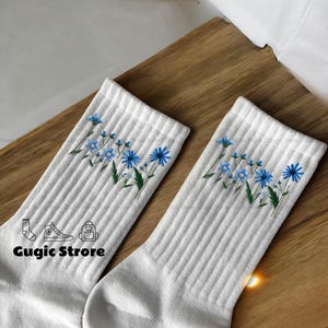 Op de afbeelding: Een paar witte sokken met blauwe bloemenborduursels. De sokken hebben een geribbelde textuur en bevatten een kleine afbeelding van een schoen, een sok en een rugzak.