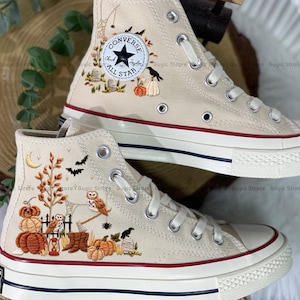 May include: Beige high-top Converse sneakers with embroidered Halloween-themed designs. The designs include pumpkins, bats, owls, and a crescent moon. The sneakers have white laces, a white rubber sole, and a red and navy blue stripe.