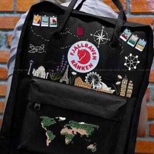 Puede incluir: Mochila negra con parches bordados con temática de viajes, incluyendo un mapa del mundo, monumentos y una brújula. Cuenta con un parche con el logo de un zorro rojo y parches tipo foto. La mochila tiene asas negras y un bolsillo frontal.