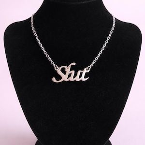 Puede incluir: Un collar plateado con la palabra "Shut" en cursiva. El colgante cuelga de una delicada cadena, expuesto sobre un busto de terciopelo negro. El collar es una pieza de joyería sencilla pero audaz.