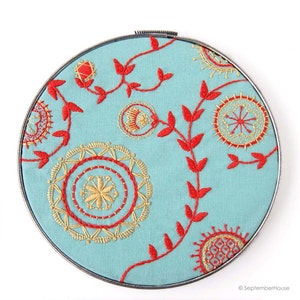 Puede incluir: Un aro de bordado turquesa con bordado floral rojo y dorado. El diseño presenta un patrón repetitivo de hojas y círculos.