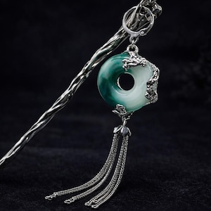 May include: A silver hair stick with a green and white jade pendant. The pendant is a circular shape with a hole in the center and silver detailing. Silver chains hang from the bottom of the pendant. The background is black.