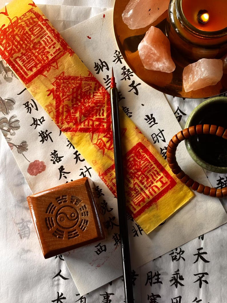 May include: Overhead view of spiritual items. A wooden block with a yin-yang symbol, a calligraphy brush, and a yellow and red paper are on a white surface with Chinese characters. A candle, crystals, and a beaded bracelet are also present.