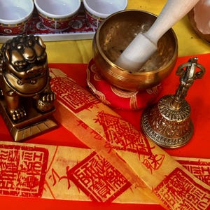 May include: A selection of spiritual items, including a golden singing bowl with a wooden mallet, a small bell, and a golden lion statue. There are also small white bowls with red and gold trim, and yellow paper with red stamps.