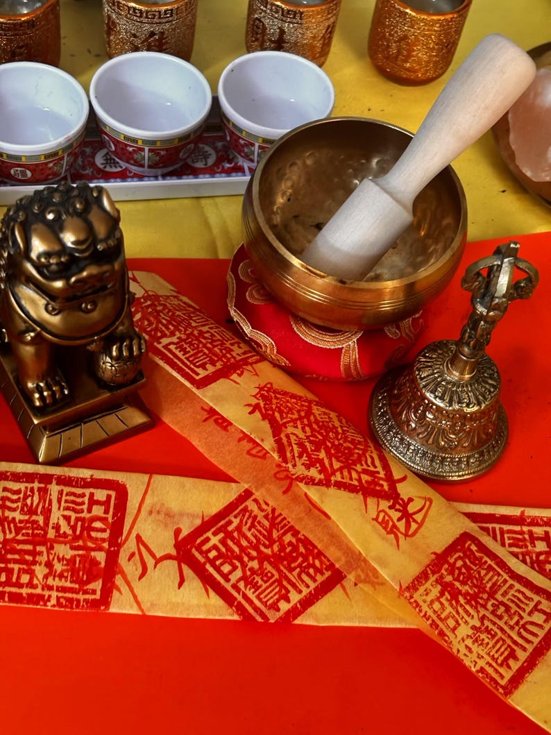 May include: A selection of spiritual items, including a golden singing bowl with a wooden mallet, a small bell, and a golden lion statue. There are also small white bowls with red and gold trim, and yellow paper with red stamps.