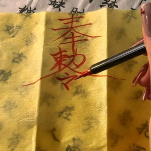 May include: A hand writing Chinese calligraphy with a red pen on yellow paper. The paper has Chinese characters printed on it in black ink. The calligraphy is in red ink.