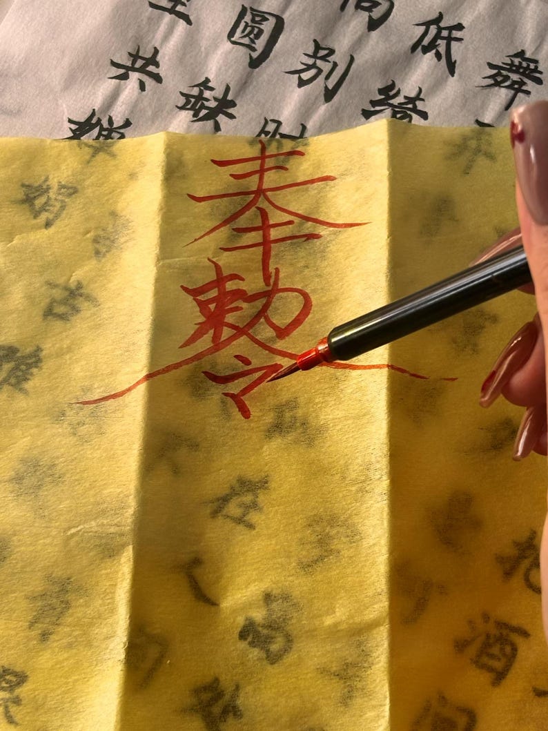 May include: A hand writing Chinese calligraphy with a red pen on yellow paper. The paper has Chinese characters printed on it in black ink. The calligraphy is in red ink.