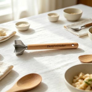 May include: A kitchen tool with a light wood handle and a silver metal crushing head. The handle features the engraved text Daisy's Crusher and a small circular hole at the end. The metal head has a multi-bladed design for crushing food items.