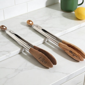 May include: Two stainless steel and wood kitchen tongs with the names "Ellen" and "Megan" engraved on the wooden handles. The tongs are resting on a white marble countertop. A green mug and a lemon are in the background.