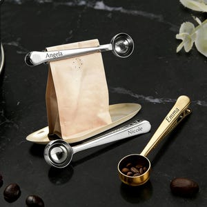 May include: A set of personalized coffee scoops with bag clips. One silver scoop with "Angela" engraved, another with "Nicole", and a gold scoop with "Emma". A brown paper bag sits in a gold tray, with coffee beans scattered around.
