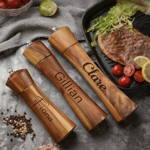 May include: Three wooden pepper grinders with silver tops, each engraved with a name: Fiona, Gillian, and Clare. The grinders are displayed on a gray surface, alongside peppercorns, tomatoes, and a steak.