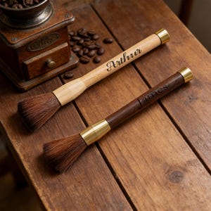 May include: Two coffee brushes with wooden handles and brass accents on a wooden surface. One is light-coloured with "Arthur" engraved, the other is dark with "James". Coffee beans and a grinder are in the background.