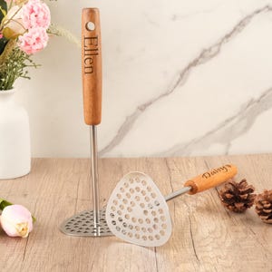 May include: Two potato mashers with wooden handles. One masher has the name "Ellen" engraved on the handle. The other masher has the name "Daisy" engraved on the handle. The mashers have a stainless steel base.