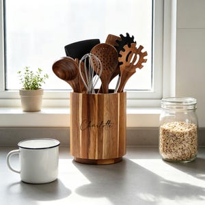 May include: A cylindrical wooden utensil holder with vertical grain patterns and the name Charlotte engraved on the front. It contains a collection of wooden spoons, a metal whisk, a black spatula, and a wooden pasta server for kitchen storage.