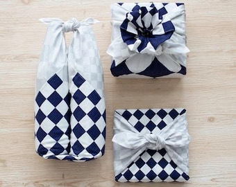 May include: Three gifts wrapped in white fabric with a blue and white checkered pattern. The fabric is tied with a knot and the ends are left loose. The gifts are arranged on a light wood surface.