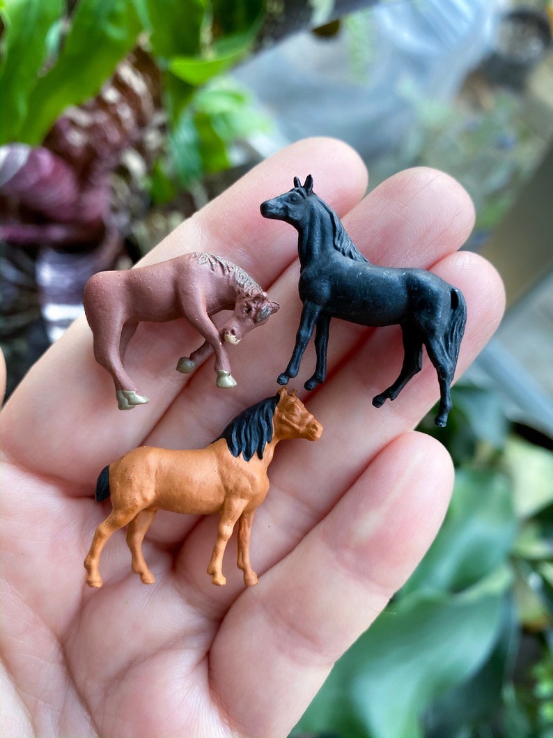 May include: Three miniature plastic horses in different colors: brown, black, and orange. The horses are standing on a hand.