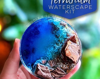 May include: A small, round terrarium with a blue resin water scene. The terrarium features a rocky shore with brown and white rocks and a blue and green water effect. The terrarium is labelled "Doodle Bird Terrariums" and "Terrarium Waterscape Kit".