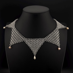 May include: A silver chainmail necklace with a triangular design. The necklace features a series of interconnected metal rings, with small pearl accents at the points and a single teardrop pearl pendant.