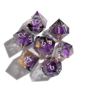 Peut inclure: Ensemble de sept dés violets et argentés avec des paillettes dorées. Les dés sont transparents et montrent les chiffres 1, 2, 3, 4, 6, 8 et 12.