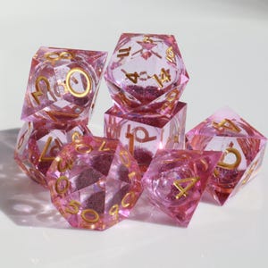 Könnte beinhalten: Ein Satz rosa Würfel mit goldenem Glitzer und goldenen Zahlen. Die Würfel sind aus klarem Harz gefertigt und haben verschiedene Formen, darunter ein W4, W6, W8, W10, W12 und W20.