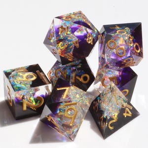 Peut inclure: Un ensemble de sept dés polyédriques. Les dés sont un mélange de résine transparente et noire, avec des chiffres dorés et des inclusions irisées. Les dés sont disposés sur une surface blanche.