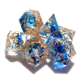 Könnte beinhalten: Satz blauer und goldener Würfel mit goldenen Flocken und Zahlen. Die Würfel sind aus klarem Harz gefertigt und haben ein wirbelndes Muster.