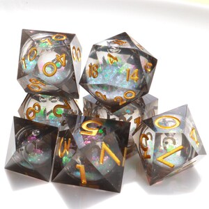 Peut inclure: Ensemble de sept dés polyédriques pour jeux de rôle. Les dés transparents présentent une teinte fumée, avec des paillettes irisées et de petites sphères blanches. Des chiffres dorés sont visibles sur chaque face.