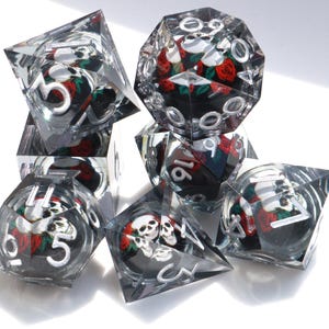 Peut inclure: Un ensemble de dés transparents à multiples faces avec un motif de crâne et de roses. Les dés ont des chiffres argentés et un fond noir avec des roses rouges et des crânes blancs. Pour les jeux de société.