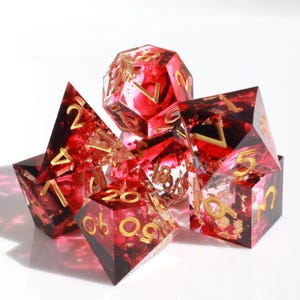 Könnte beinhalten: Satz von sieben roten und goldenen Würfeln mit goldenen Flocken und Zahlen. Die Würfel sind aus klarem Harz gefertigt und haben verschiedene Formen, darunter ein W20, W12, W10, W8, W6 und W4.