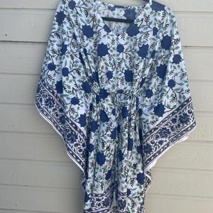 Puede incluir: Un caftán blanco con un estampado floral azul y hojas verdes, colgado de una percha negra. El caftán tiene cuello en V y cintura con cordón. Los bordes están adornados con un borde floral azul.