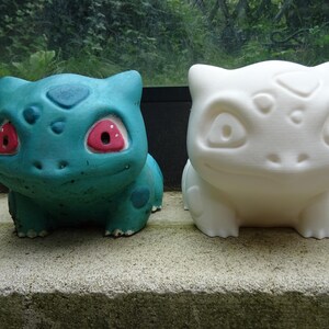 Peut inclure: Deux figurines en céramique d'un personnage de Pokemon, l'une peinte en bleu turquoise avec des yeux rouges et l'autre peinte en blanc.