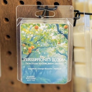 May include: A clear plastic package containing scented wax cubes. The label features a watercolor illustration of a tree with orange fruit. Text on the label reads "PERSEPHONE'S BLOOM" and lists scent notes. The brand name "SASSE" is also visible.
