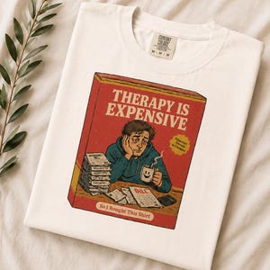 Puede incluir: Una camiseta de color crema con un gráfico de una persona sentada en un escritorio con una pila de facturas y una taza. El texto dice "THERAPY IS EXPENSIVE", "Financial Therapy is Cheaper" y "So I Bought This Shirt". La camiseta tiene una etiqueta Comfort Colors.