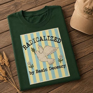 May include: A dark green t-shirt featuring a graphic with vertical light blue and yellow stripes, a white dove holding an olive branch, two small flowers, and the text "RADICALIZED by Basic Decency" in black, accompanied by a tan baseball cap.