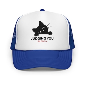May include: A blue and white trucker hat featuring a graphic of a black cat peeking over the edge. The text JUDGING YOU is printed in black capital letters, with the word SILENTLY printed in red capital letters directly underneath the cat graphic.