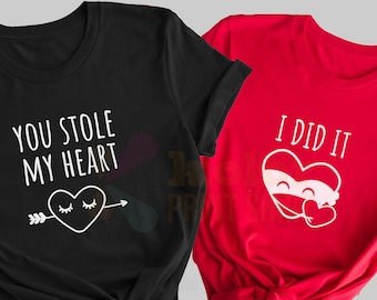 Puede incluir: Dos camisetas, una negra y otra roja, con un diseño de pareja a juego. La camiseta negra dice "You stole my heart" con un gráfico de corazón y flecha. La camiseta roja dice "I did it" con un gráfico de corazón.