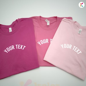 May include: Three folded t-shirts in shades of pink, from deep magenta to light pink. Each shirt has the text "YOUR TEXT" printed in white across the front. The shirts are neatly arranged on a white surface.